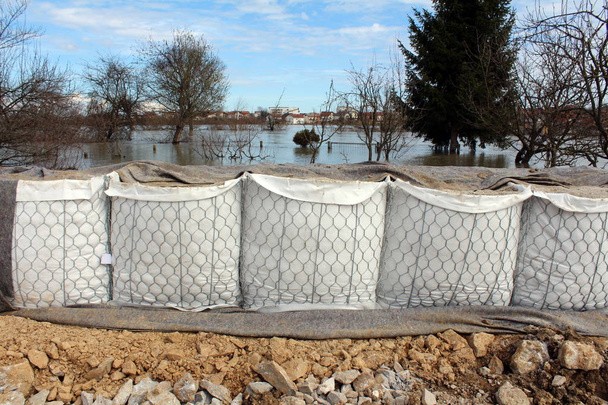 Novas barreiras de caixa de areia proteção contra inundações parcialmente cobertas com tecido geotêxtil, rochas maciças na frente e rio inundado, árvores e casas no fundo
 - Foto, Imagem