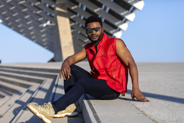 Front view of young black man wearing sunglasses sitting on staircase in a sunny day while looking camera - Foto, afbeelding