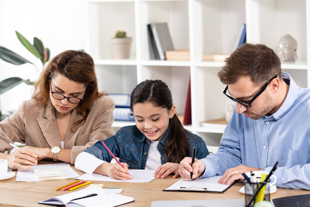 bambino felice che disegna vicino a genitori in occhiali che lavorano in ufficio
  - Foto, immagini