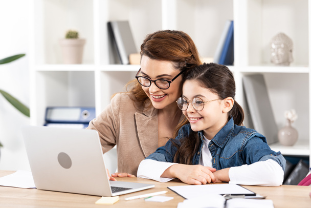 cheerful mother sitting with cute daughter in glasses and looking at laptop  - Fotografie, Obrázek