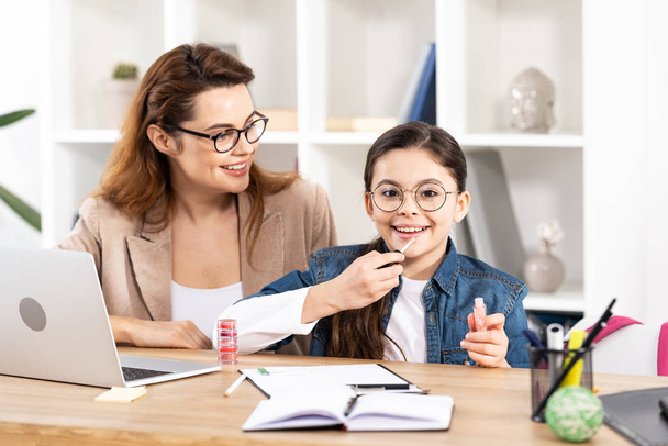 cheerful mother looking at cute daughter applying lip gloss in office  - Fotó, kép