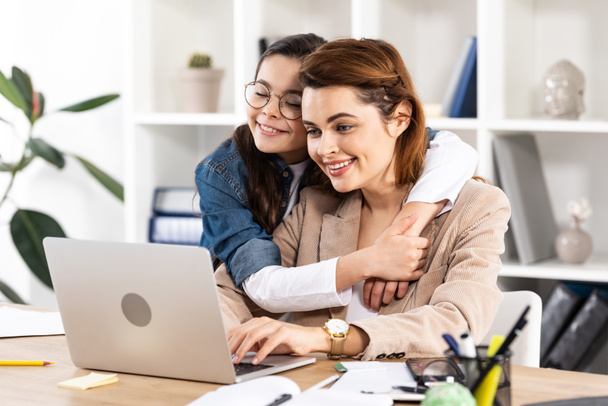 happy daughter hugging cheerful mother using laptop in office  - Photo, Image