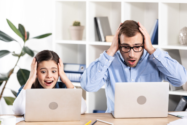 σοκαρισμένος πατέρας και κόρη κοιτάζοντας φορητούς υπολογιστές και κρατώντας το κεφάλι στο γραφείο  - Φωτογραφία, εικόνα
