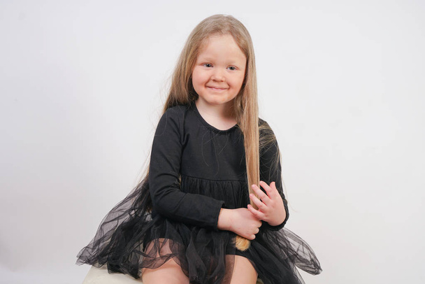 niedliche kleine Mädchen mit langen blonden Haaren sitzt und berührt ihre Haare auf weißem Hintergrund - Foto, Bild