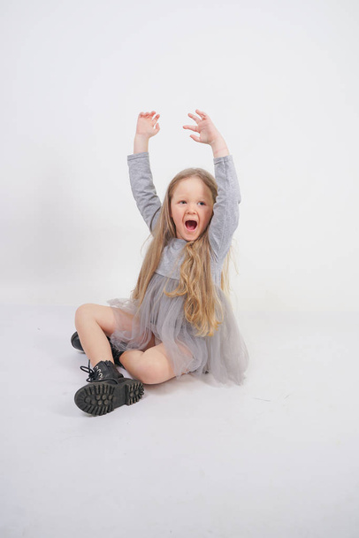 cute girl child with long blond hair sitting on the floor and yawns sweetly, stretching her hands in different directions on white studio background - Zdjęcie, obraz