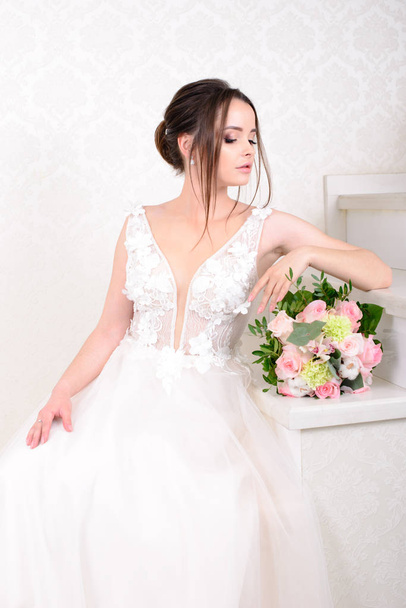 Štíhlá krásná žena na sobě luxusní svatební šaty přes bílé studio pozadí. Nádherná nevěsta drží květiny. - Fotografie, Obrázek