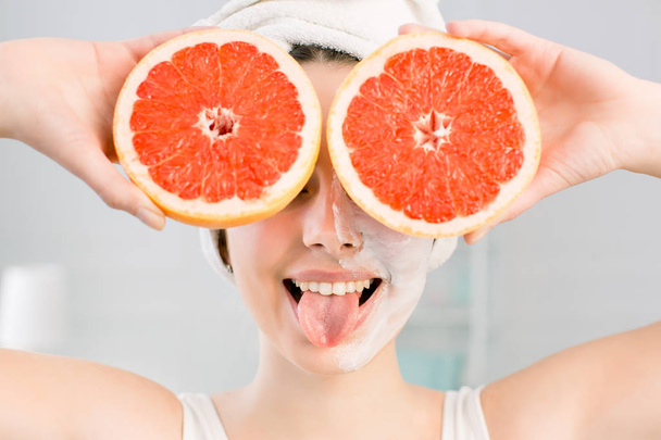 Young cheerful european woman with white facial mask and white towel on head shows tongue and holds big grapefruits, covering her eyes. Healthy diet, skincare, facial treatment, cosmetology concept. - Fotó, kép