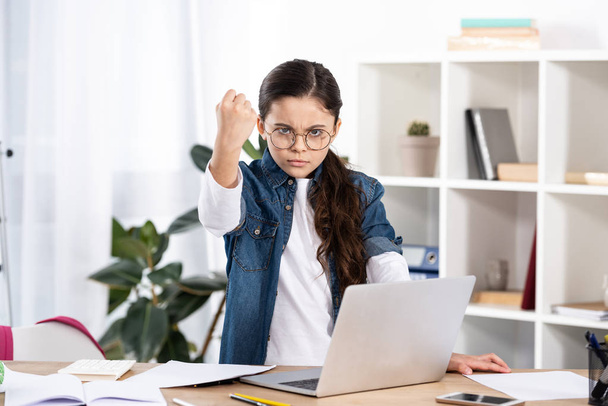 cute kid in glasses showing fist while standing near laptop in office  - Foto, afbeelding
