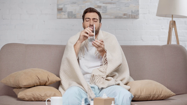 schöner kranker Mann sitzt auf Sofa unter Decke, nimmt Flasche mit Tabletten, trinkt Wasser und niest im Wohnzimmer  - Filmmaterial, Video