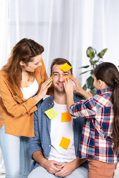cheerful woman and kid putting yellow sticky notes on handsome man  - Foto, afbeelding