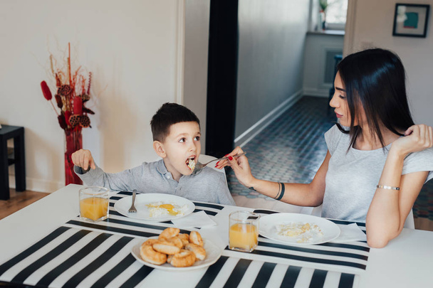 Beautiful young woman at home with her little cute son are having breakfast. - Photo, Image
