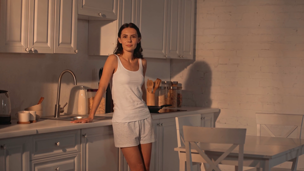 ελκυστική γυναίκα με σφικτά χέρια στέκεται με το χέρι στο ισχίο, χαμογελώντας και κοιτάζοντας την κάμερα στην κουζίνα με ηλιακό φως  - Πλάνα, βίντεο