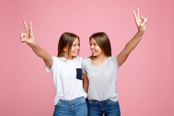 Pretty two young twin sisters with beautiful smile with hands up showing peace gesture over pink background. - Fotografie, Obrázek