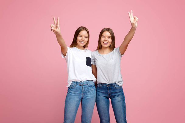 Image of charming two young twin sisters with beautiful smile with hands up showing peace gesture over pink background. - Fotografie, Obrázek