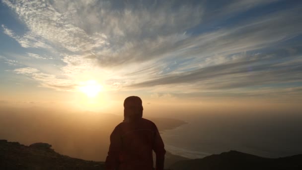 La caméra suit un homme approchant un bord de falaise sous le vent fort au-dessus de l’océan. L’homme lève les mains vers le haut au beau coucher du soleil au ralenti. - Séquence, vidéo