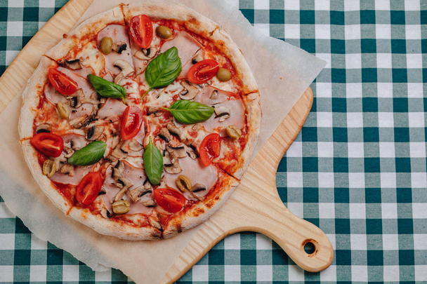 Napolilainen pizza kinkulla, juustolla, arugulalla, basilikalla, tomaateilla, jotka on siroteltu juustolla puulaudalle pöytäliinalla sellissä, jossa on tekstipaikka
 - Valokuva, kuva