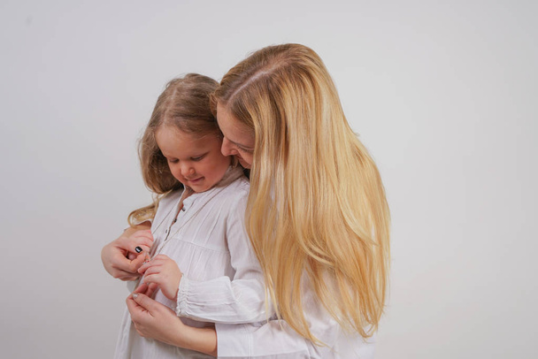 anya és lánya, fehér ing, hosszú szőke haja jelentő szilárd hátteret a stúdióban. bájos család gondoskodik egymásra, és megölel. - Fotó, kép