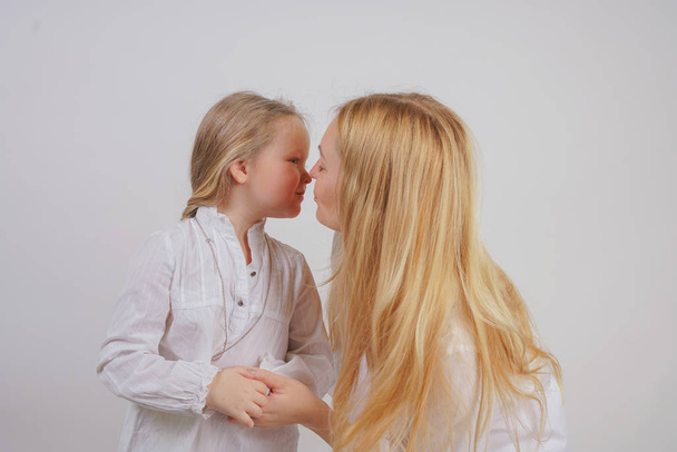 mom and daughter in white shirts with long blonde hair posing on a solid background in the Studio. charming family takes care of each other and hugs. - Fotografie, Obrázek