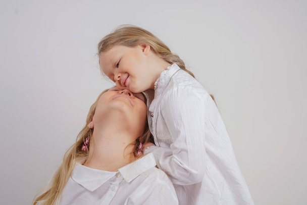 mãe e filha em camisas brancas com cabelo loiro longo posando em um fundo sólido no estúdio. uma família encantadora cuida uns dos outros e faz tranças
. - Foto, Imagem