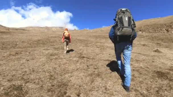 Δύο φωτογράφοι τουριστών με σακίδια σε καπέλα και γυαλιά ηλίου πηγαίνουν στο λόφο στο κίτρινο γρασίδι με κάμερες στα χέρια τους στο φόντο των χιονισμένων βουνών. αργή κίνηση - Πλάνα, βίντεο