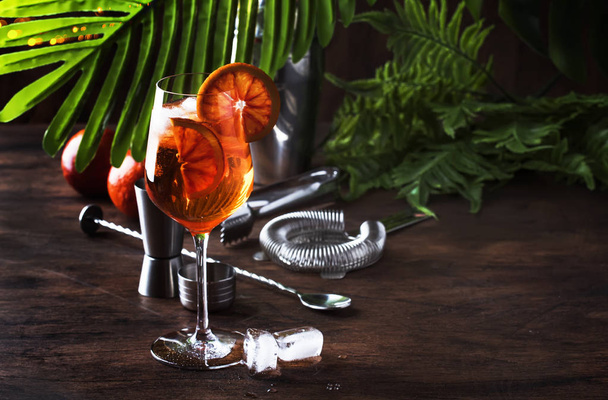Aperol spritz cocktail in big wine glass with orange slices, summer cool fresh alcoholic cold beverage. Wooden bar counter background with tools, summer mood concept with palm leaves, copy space, selective focus - Zdjęcie, obraz