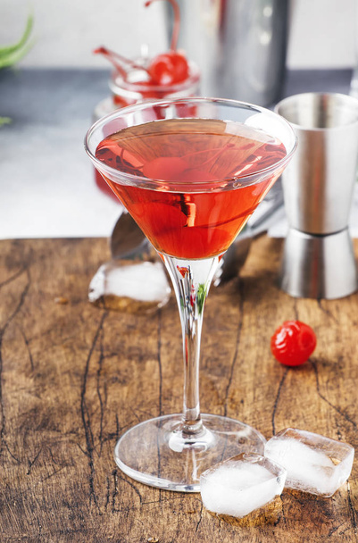 Classic alcoholic cocktail Manhattan with american bourbon, red vermouth, bitter, ice and cocktail cherry in glass, tools, gray bar counter background, copy space, selective focus - Fotó, kép
