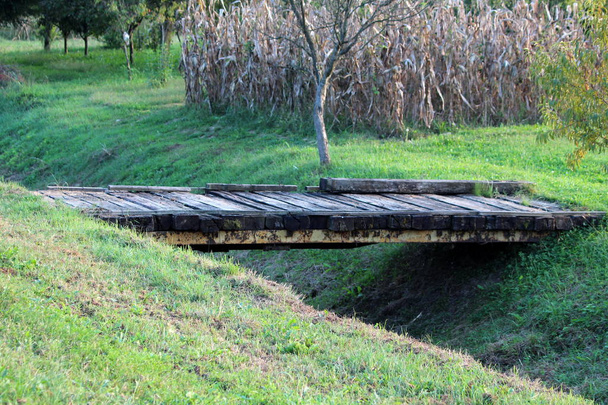 kurze hausgemachte Holzbrücke mit starken Metallträgern, die zwei Seiten des kleinen Kanals hinter lokalen Gärten mit Bäumen und Maisfeld an einem warmen, sonnigen Tag bei Sonnenuntergang verbindet - Foto, Bild