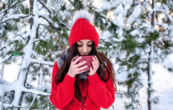 Young brunette woman in red hat drinking coffee in snowy winter park - Photo, Image