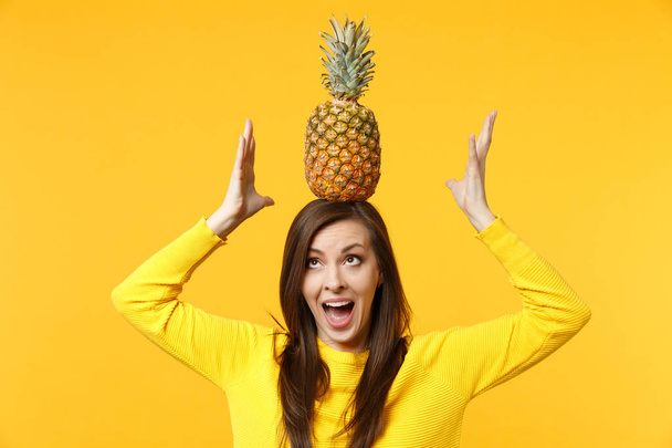 Veselá, vzrušená mladá žena v příležitostných šatech s čerstvým zralým ananasovým ovocem na hlavě izolovaná na žlutém oranžovém pozadí. Lidé mají živý životní styl, relaxační koncept. Vysmívat prostor pro kopírování. - Fotografie, Obrázek