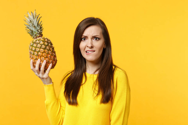 Preoccupied nervous young woman in casual clothes biting lips, hold fresh ripe pineapple fruit isolated on yellow orange background. People vivid lifestyle, relax vacation concept. Mock up copy space. - Photo, Image