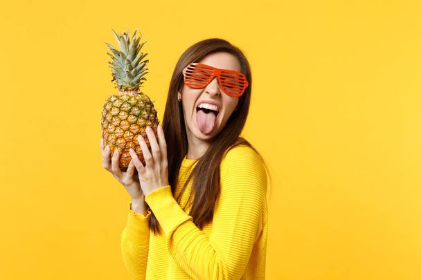 Crazy young woman in funny glasses showing tongue, holding in hands fresh ripe pineapple fruit isolated on yellow orange background. People vivid lifestyle relax vacation concept. Mock up copy space. - Photo, Image