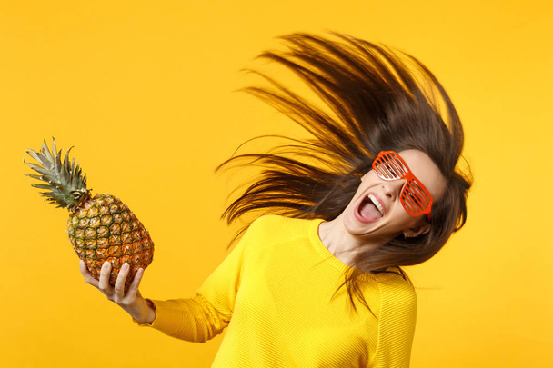 Cheerful young woman in funny glasses with flowing hair holding fresh ripe pineapple fruit isolated on yellow orange wall background. People vivid lifestyle relax vacation concept. Mock up copy space. - Photo, Image