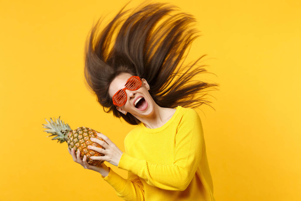 Excited young woman in funny glasses with flying hair holding fresh ripe pineapple fruit isolated on yellow orange wall background. People vivid lifestyle, relax vacation concept. Mock up copy space. - Photo, Image