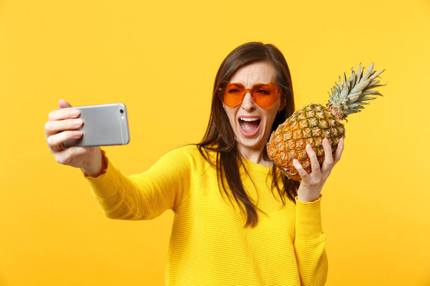 Płacz krzyczeć młoda kobieta w okularach trzymać owoc ananasa robi selfie strzał na telefon komórkowy izolowane na żółtym pomarańczowym tle. Ludzie żywy styl życia relaks koncepcja wakacje. Makiety przestrzeni kopiowania. - Zdjęcie, obraz
