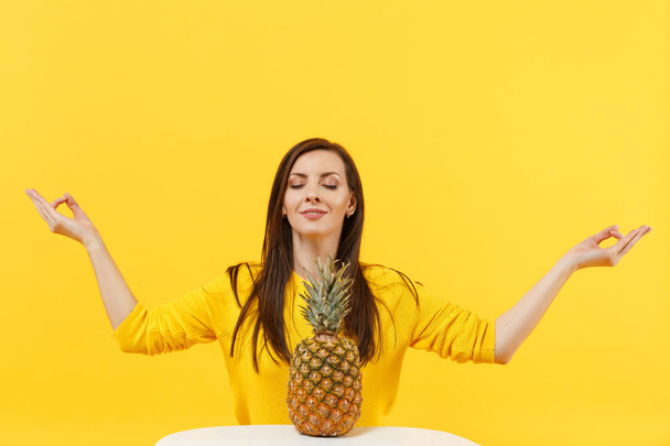 Smiling young woman with fresh pineapple fruit keeping eyes closed hold hands in yoga gesture, relaxing meditating isolated on yellow orange background. People vivid lifestyle, relax vacation concept. - Photo, Image