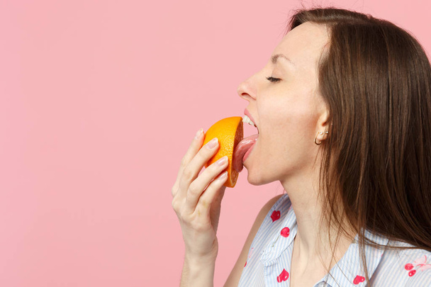 Side view of young woman in summer clothes keeping eyes closed licking half of fresh orange fruit isolated on pink pastel background. People vivid lifestyle relax vacation concept. Mock up copy space. - Φωτογραφία, εικόνα