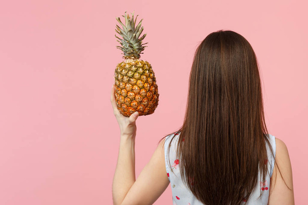 Rückansicht einer jungen Frau in Sommerkleidung, die frische reife Ananasfrüchte in der Hand hält, isoliert auf rosa Pastell-Hintergrund. Menschen lebendigen Lebensstil, entspannen Urlaubskonzept. Attrappe Kopierraum. - Foto, Bild