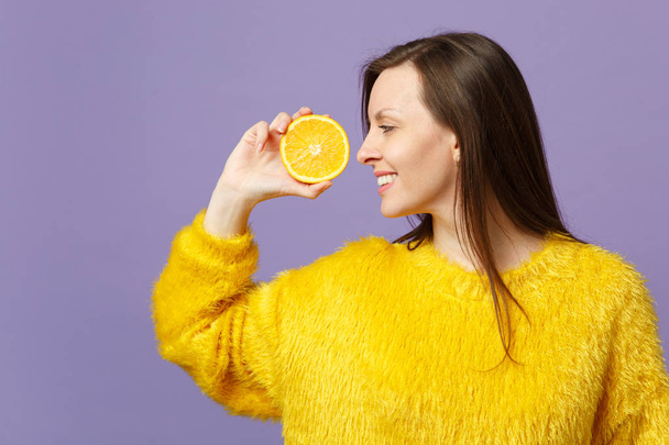 Smiling young woman in fur sweater hold, looking aside on half of fresh ripe orange fruit isolated on violet pastel wall background. People vivid lifestyle, relax vacation concept. Mock up copy space. - Photo, Image