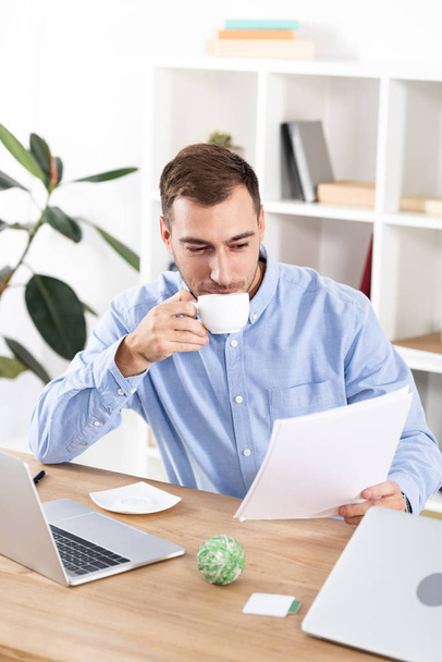 cheerful businessman drinking coffee while sitting in office and holding blank papers - Фото, изображение