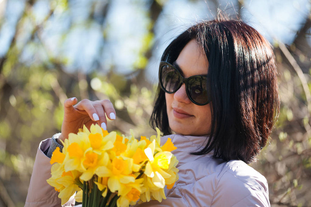 Bir buket Daffodils tutan güneş gözlüğü genç güzel esmer kız. Bir avuç çiçek - Fotoğraf, Görsel