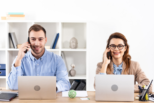 successful businessman and businesswoman talking on smartphones near laptops in office  - Фото, зображення