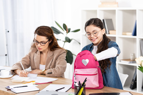 cheerful kid looking at backpack near mother in glasses working in office  - 写真・画像