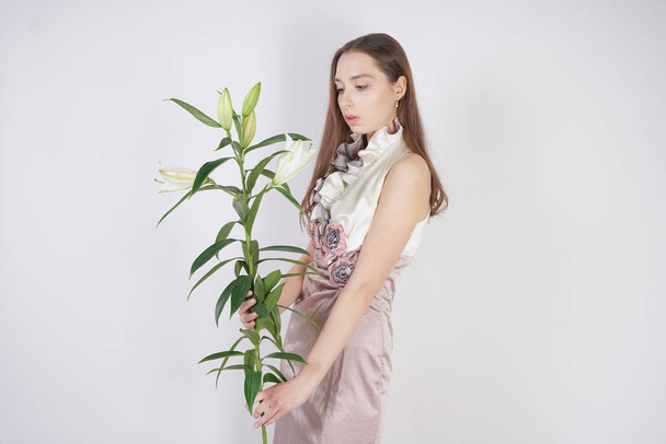 charming young caucasian girl in a pretty dress holds a lush Lily flower in her hands and stands on a white background in the Studio alone - Foto, imagen