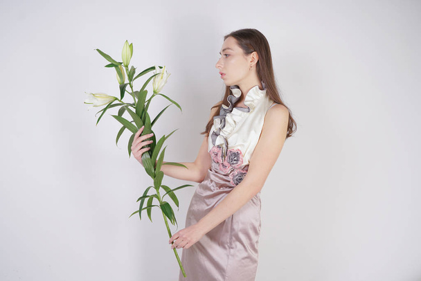charming young caucasian girl in a pretty dress holds a lush Lily flower in her hands and stands on a white background in the Studio alone - Photo, Image