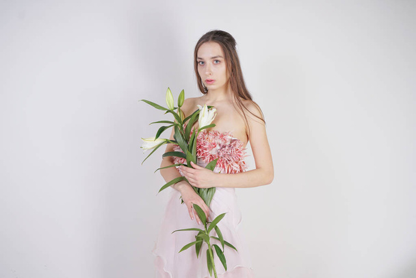 charming young caucasian girl in a pretty dress holds a lush Lily flower in her hands and stands on a white background in the Studio alone - Photo, image