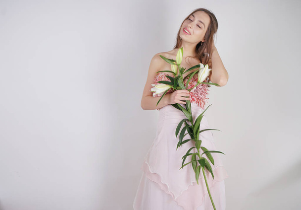 charming young caucasian girl in a pretty dress holds a lush Lily flower in her hands and stands on a white background in the Studio alone - Photo, Image
