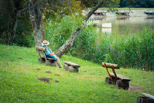 Male farmer sitting on wooden bench close to pond  - 写真・画像