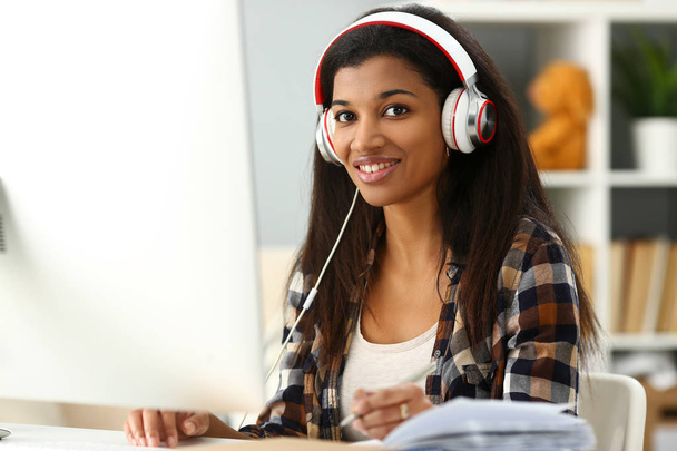 Mujer sonriente negra sentada en el lugar de trabajo usando auriculares
 - Foto, Imagen