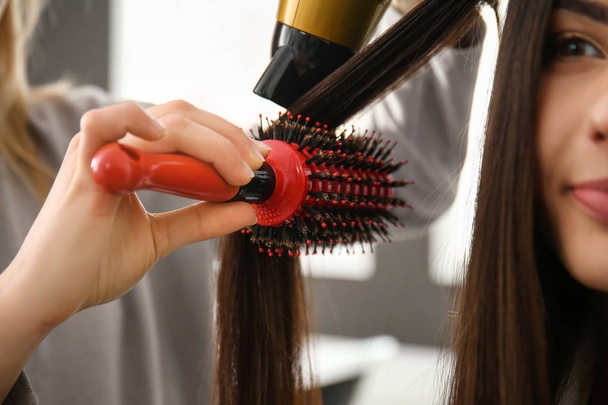 Hairdresser with blow dryer brushing long hair of young woman in salon - Fotó, kép