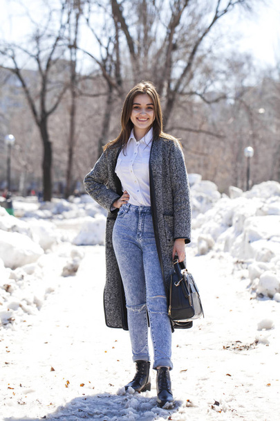Brunette girl in a gray coat and blue jeans walks in spring park - Photo, Image